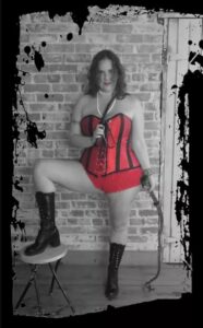 Gaia Morissette in a red corset holding a whip and riding crop